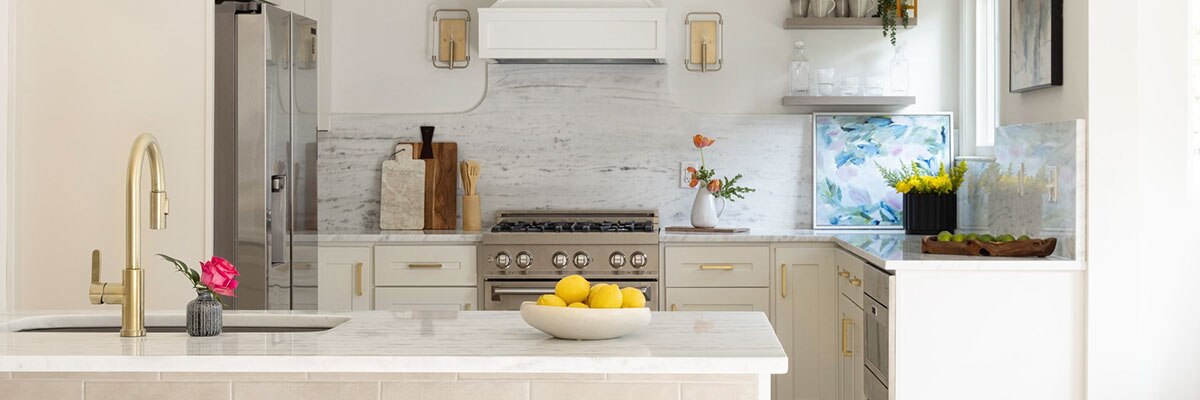 Renovated kitchen with gray marble backsplash and countertops, island base covered by beige subway tile, off-white cabinets with brass accents.