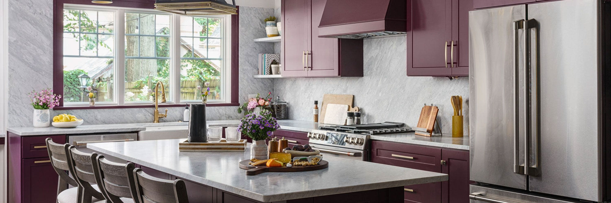 Renovated kitchen with plum-colored cabinets, gray marble backsplash and countertops, stainless steel appliances, and farm sink in front of three windows.