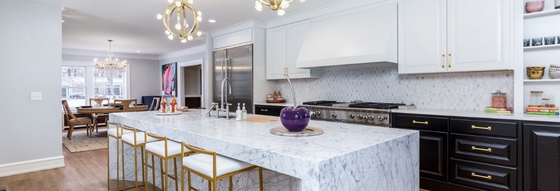 Kitchen with marble mosaic backsplash, white quartz countertops, marble waterfall island with sink, white cabinets, and brushed brass pendant lights.