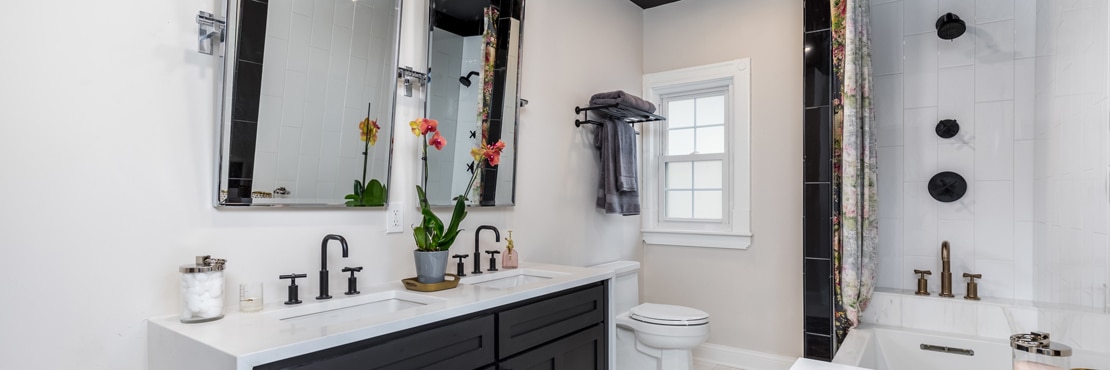 Remodeled bathroom with glossy white ceramic shower tile with glossy black shower tile accent, black shower fixtures, and vanity with double sinks.