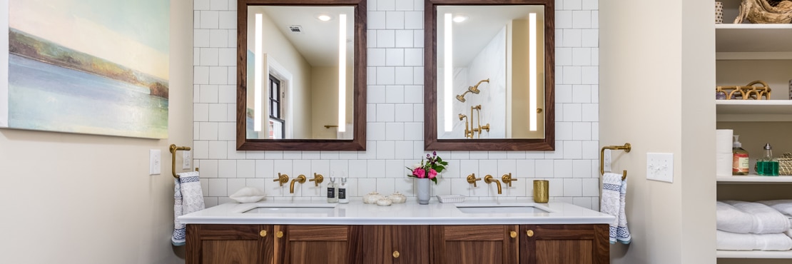 Remodeled bathroom with white square ceramic tile backsplash, natural wood vanity with dual sinks, wood framed mirrors, and brushed brass wall-mounted faucets.
