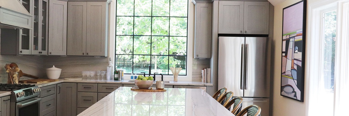 Renovated kitchen with heavily veined gray quartzite backsplash, island, and countertop, gray cabinets, stainless steel appliances, and sink in front of large picture window.