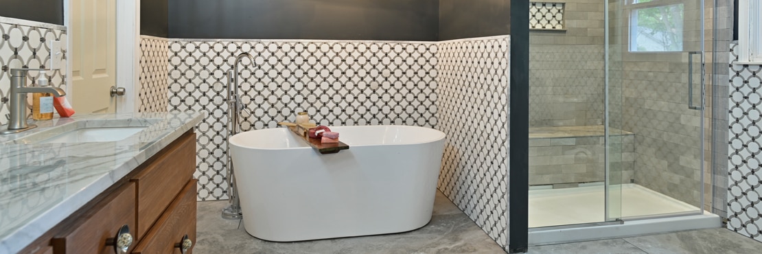 Bathroom with free standing bathtub in front of wainscoting of white marble tile with gray rings and shower with gray tile and niche with matching marble tile with rings.