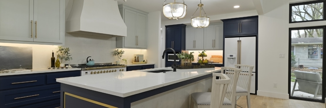 Kitchen with white upper cabinets, navy lower cabinets, white quartz countertops, island with sink, glass & brushed brass pendant lights.