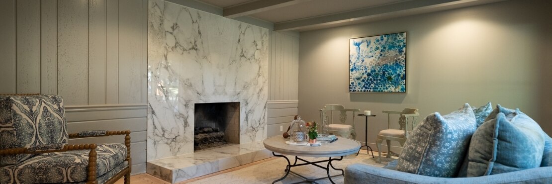 Living room with cream & gray bookmatched marble look fireplace surround and hearth, blue velvet couch, and blue & white abstract painting.