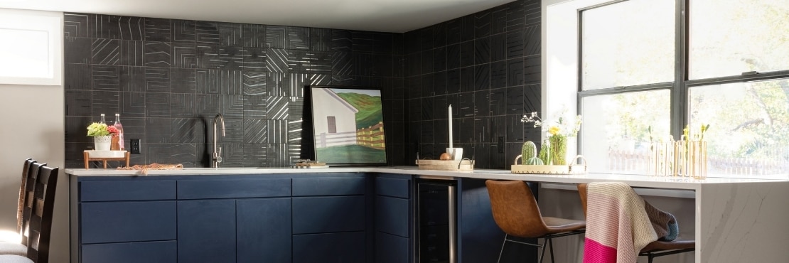Basement wet bar with black backsplash tile with geometric designs, white quartz countertop, navy cabinets, and wood floor.