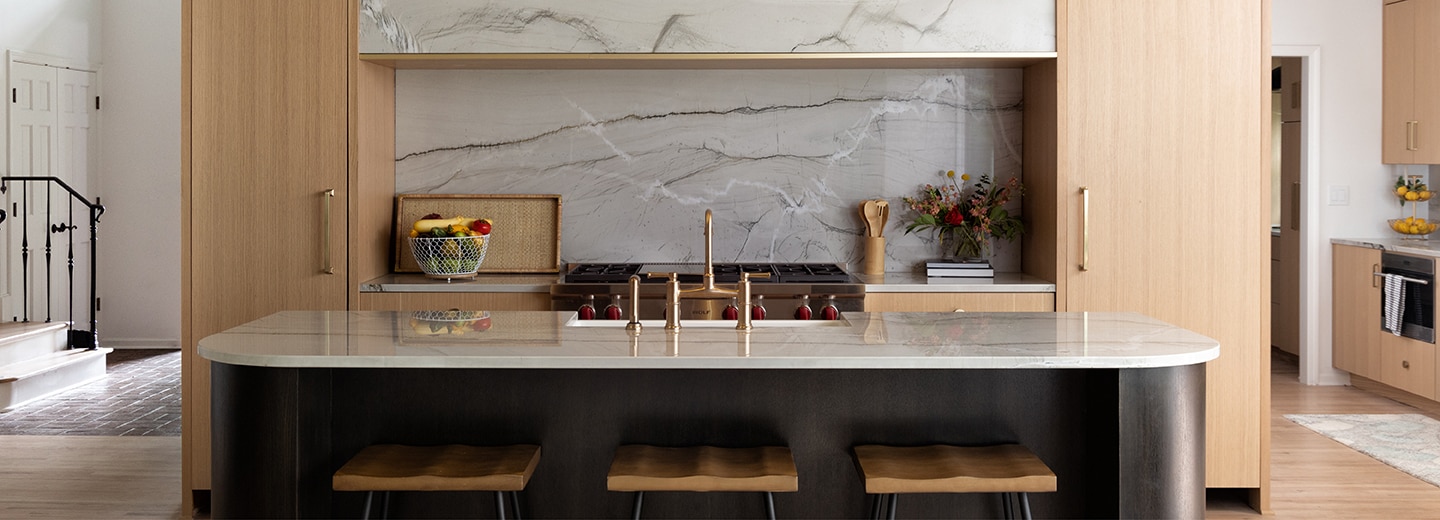 Kitchen with center island, gray quartzite countertop and backsplash, light wood cabinets, and light wood flooring.