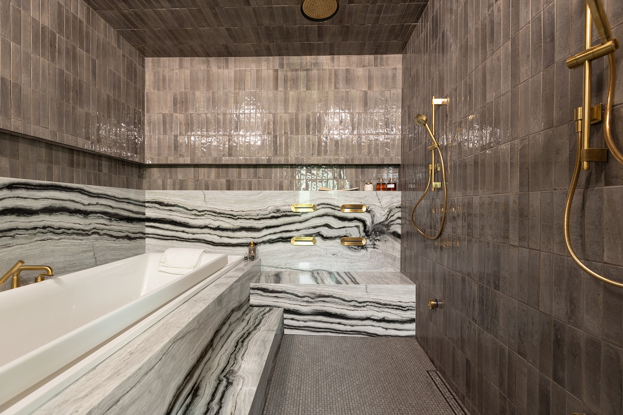 Wet room with a brick joint mosaic floor tile, gray subway tiles along walls and ceiling, and a black and white striped stone marble slab around the bath tub and seating area.