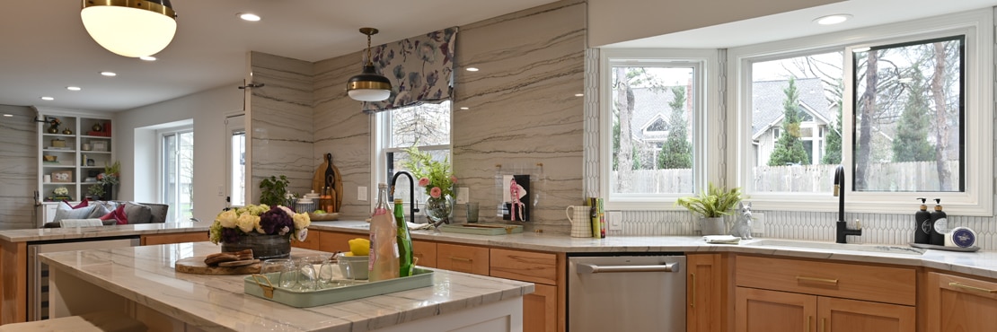Remodeled kitchen with gray heavily veined quartzite countertop, backsplash, and island, natural wood cabinets, white picket wall tile frames the bay window in front of the sink.