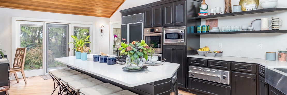 Renovated kitchen featuring white quartz countertop and island, dark wood cabinets, floating shelves, wood slat vaulted ceiling, and large picture windows.