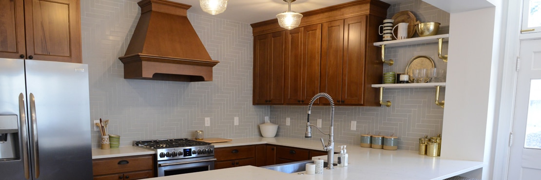 Remodeled kitchen with gray wall tile in a herringbone pattern, white quartz countertop & peninsula, natural wood cabinets & vent hood, and floating shelves.