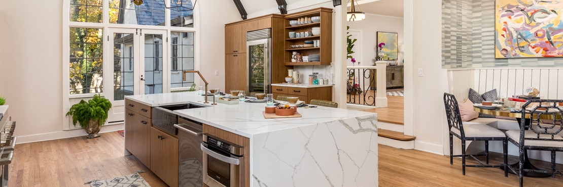 Remodeled kitchen with high vaulted ceiling, large picture windows, white & gray marble look quartz waterfall island and backsplash, wood cabinetry and floating shelves.