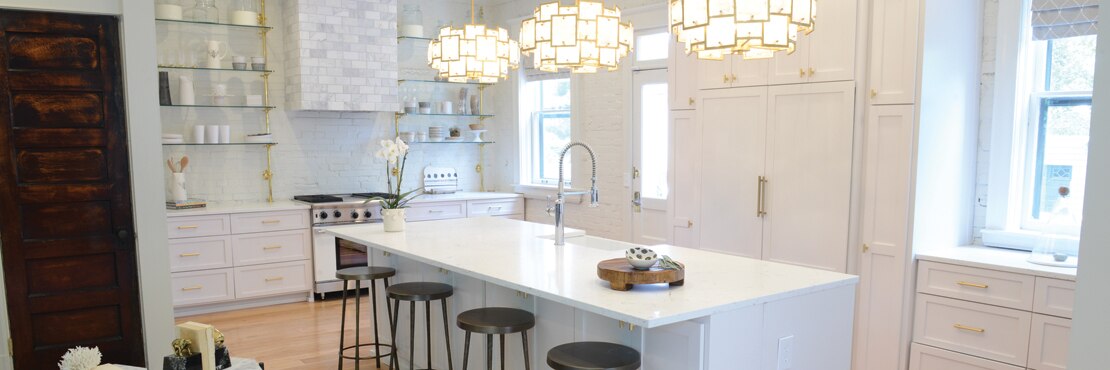 Renovated kitchen with white & gray veining quartz island with built-in sink and pendant lighting, white cabinets, floating shelves, and marble covered vent hood.