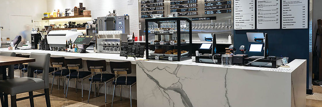 Brewery/coffee shop with white & gray marble look porcelain slab counter, stainless steel coffee/espresso machine, and wall-mounted tap beer handles.