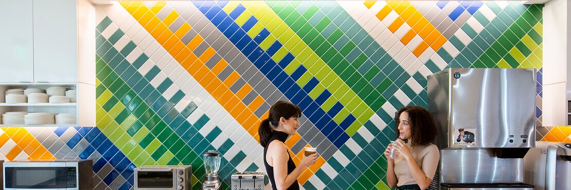 Breakroom with two women conversing in front of wall with bright green, blue, orange, and white patterned tile, white lower cabinets, and stainless steel kitchen appliances.