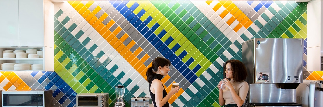 Breakroom with two women conversing in front of wall with bright green, blue, orange, and white patterned tile, white lower cabinets, and stainless steel kitchen appliances.