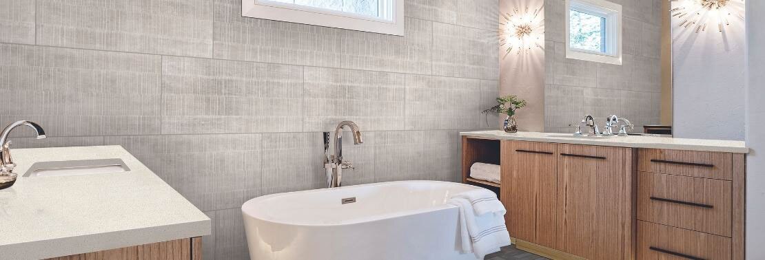 Bathroom with gray fabric look wall tile, soaker tub, separate natural wood vanities with white quartz counters, starburst wall sconces.