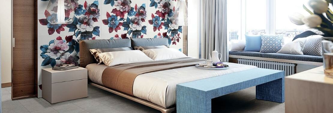 Bedroom with white slab wall tile with large white, blue, and cranberry red flowers, platform king size bed with beige & blue bedding, and blue footboard.