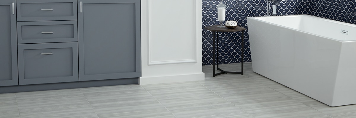 Bathroom with gray floor tile that looks like stone with linear movement, navy blue fan mosaic wall tile, chandelier over soaking bathtub, and gray-blue cabinets.