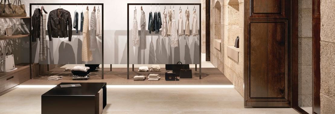 Clothing store with beige floor and wall tile that looks like limestone, racks of women’s clothing, floating wood shelves holding purses and duffle bags.