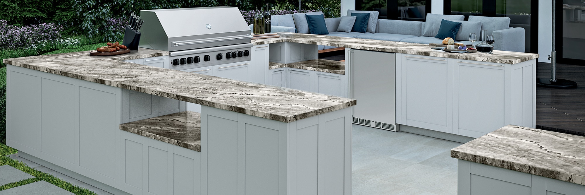 Outdoor kitchen with tan heavily veined quartzite countertops on white cabinets, stainless steel grill, gray stone-look tile flooring, and gray stone-look pavers set in grass.