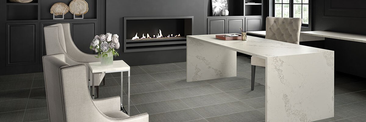 Home office with gray floor tile that looks like linen fabric, desk made of white quart with gray veining, wall-mounted fireplace, dark gray built-in and walls.