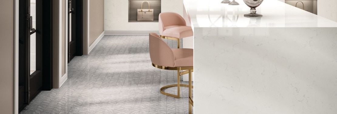 Boutique with white quartz waterfall countertop, pink & brass chairs, gray chevron mosaic floor tile, white quartz seamless wall with open shelves holding handbags.
