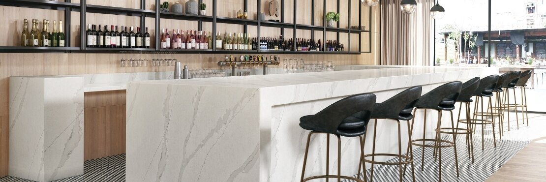 Hotel bar with white & gray marble look quartz countertops, black & white checkered tile mat set in wood look tile flooring, and racks holding wine bottles in front of wood panel wall.