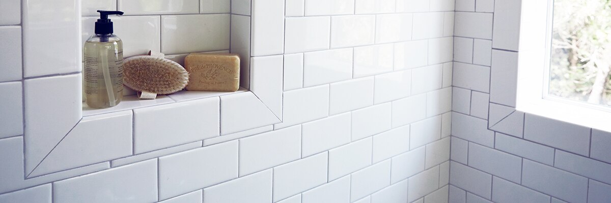 Closeup of shower wall of white subway tile, gray grout, and niche holding a soap dispenser, brush, and bar of soap.