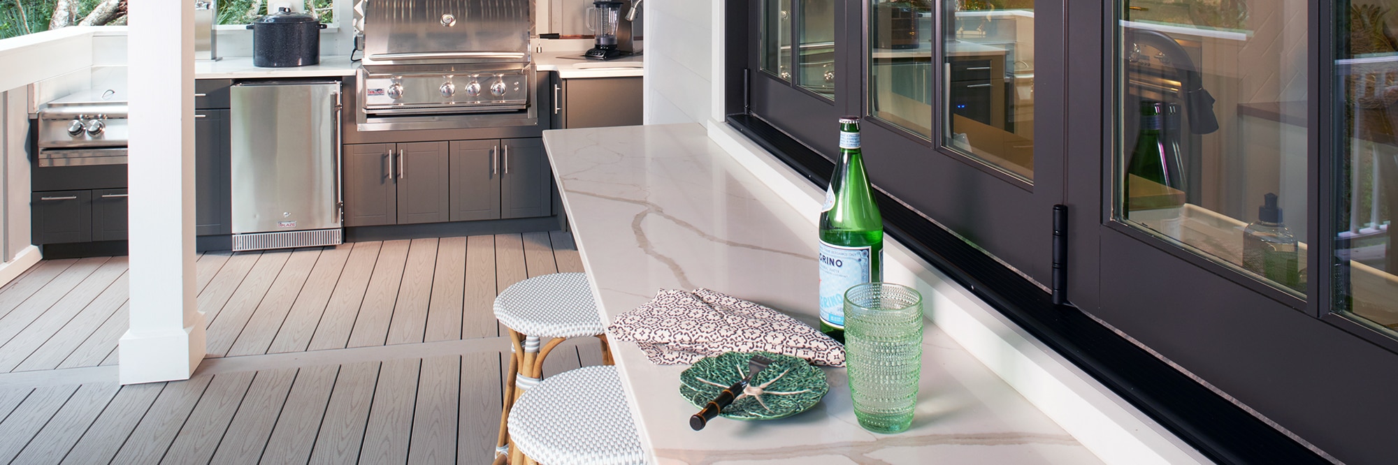 Covered patio with white quartz counter with gray veining and rattan barstools, outdoor kitchen with stainless steel grill, refrigerator, and gray cabinets.