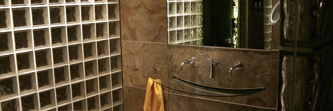 Bathroom with vanity of tan slate stone backsplash, wall-mounted glass sink, wall-mounted polished silver faucet, and glass block wall.