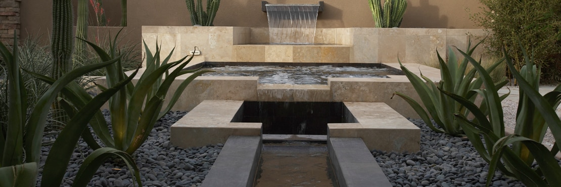 Rustic outdoor pool, spa, and fountain with tan travertine natural stone, aloe, saguaro and senita cactus in ground covered with rocks.