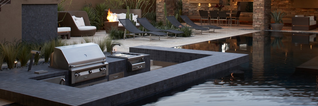 Outdoor pool with swim-up bar & grill with tan travertine natural stone deck, black lounge chairs, wicker loveseats next to firepit and aloe, saguaro and senita cactus.