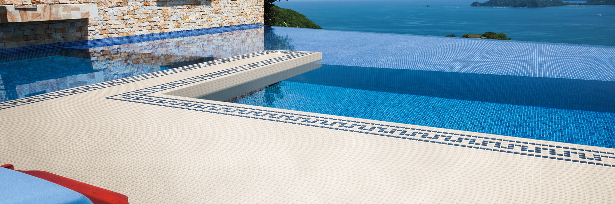 Infinity pool with blue submerged tile, white tile deck with blue border design, and stunning view of the ocean.