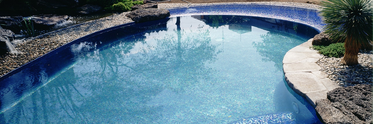 Tropical pool with iridescent blue tile, light gray paver deck, lava rocks, and palm trees set in gravel.