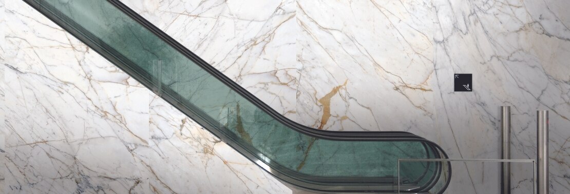 Escalator with green-tinted glass railing, large-format porcelain slab wall that looks like white marble with gold and gray veining.