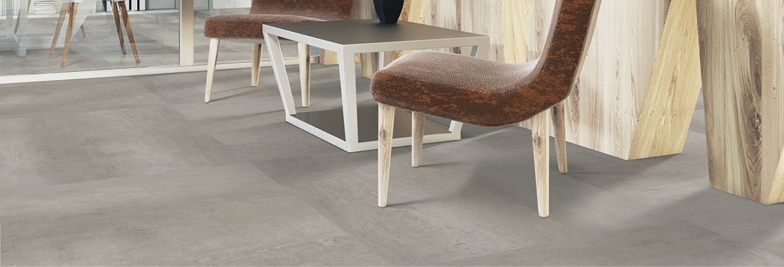 Closeup of flooring of office foyer with luxury vinyl tile that looks like gray concrete flooring, brown leather chairs and side table in front of natural wood panel wall.
