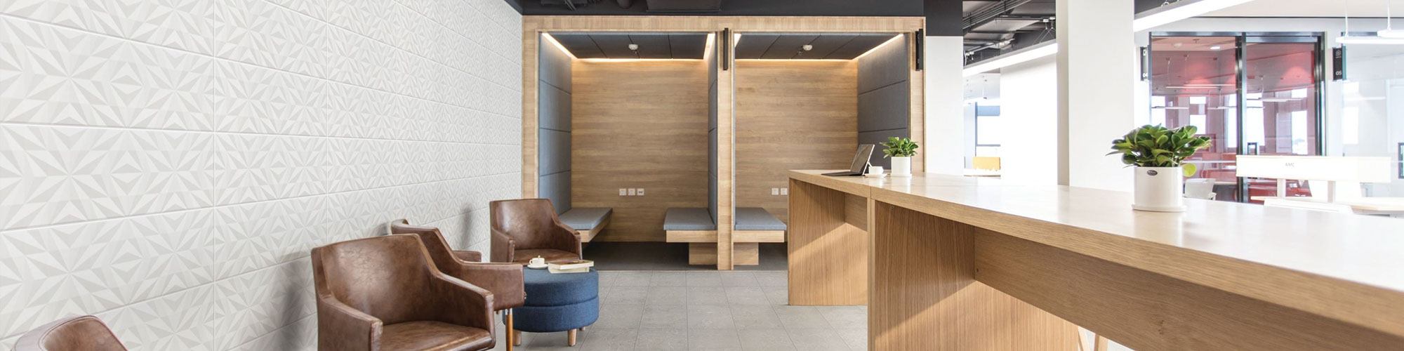 Office breakroom with floor tile that looks like gray stone, feature wall with white textured tile, brown leather chairs, long wood counter, round lighting hanging from black metal ceiling.