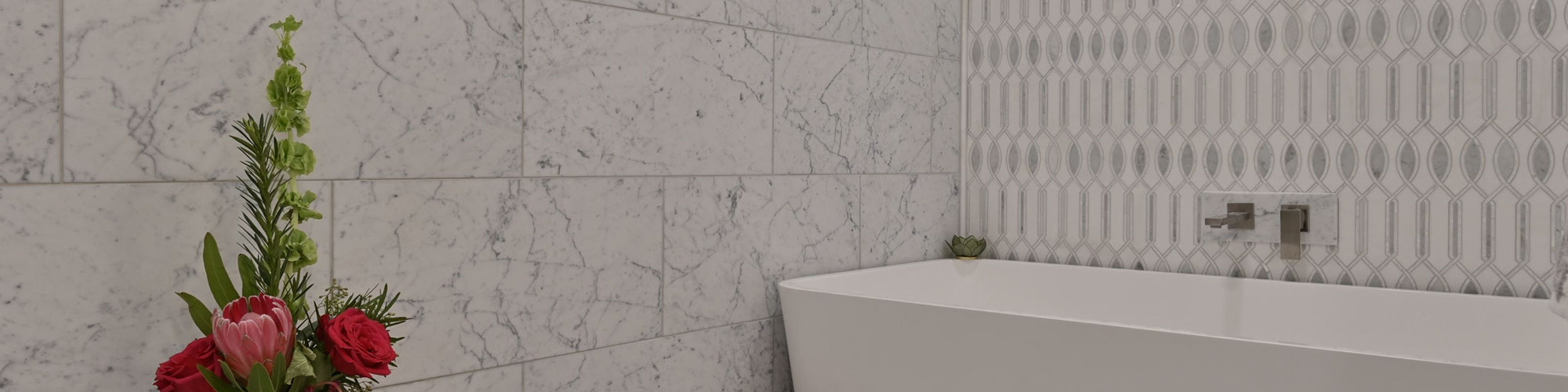 Wet room with white & gray vein marble wall tile, white & gray marble mosaic backsplash behind soaker tub, floating shelf holding bouquet of red flowers.