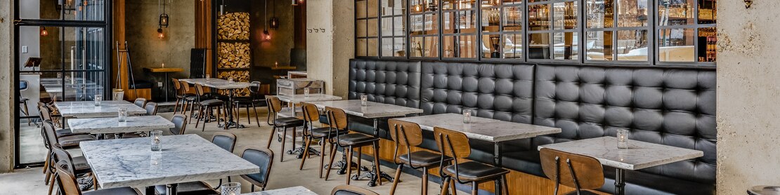 Restaurant dining room with heavy veining gray marble tabletops and wall tile, booths with tufted black leather backs, and natural wood accents.