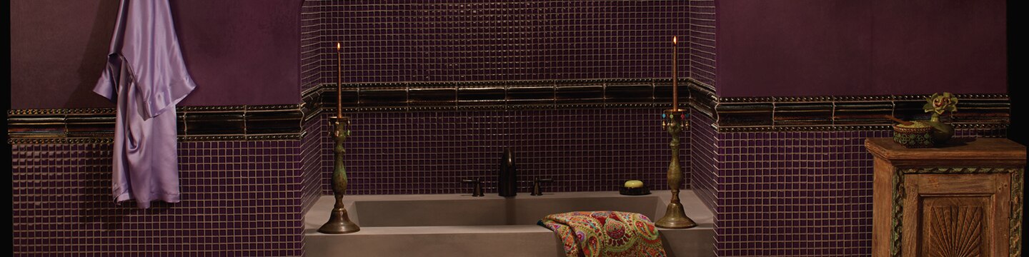 Bathroom with rich purple wallpaper, dark purple trim over wainscot of purple glass mosaic tile, and tan soaking tub with tall candlesticks.