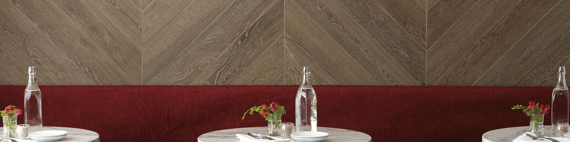 Restaurant dining room with wall-length booth covered in red velvet, herringbone wood look wall tile, marble tables with place setting & red flowers in vase.
