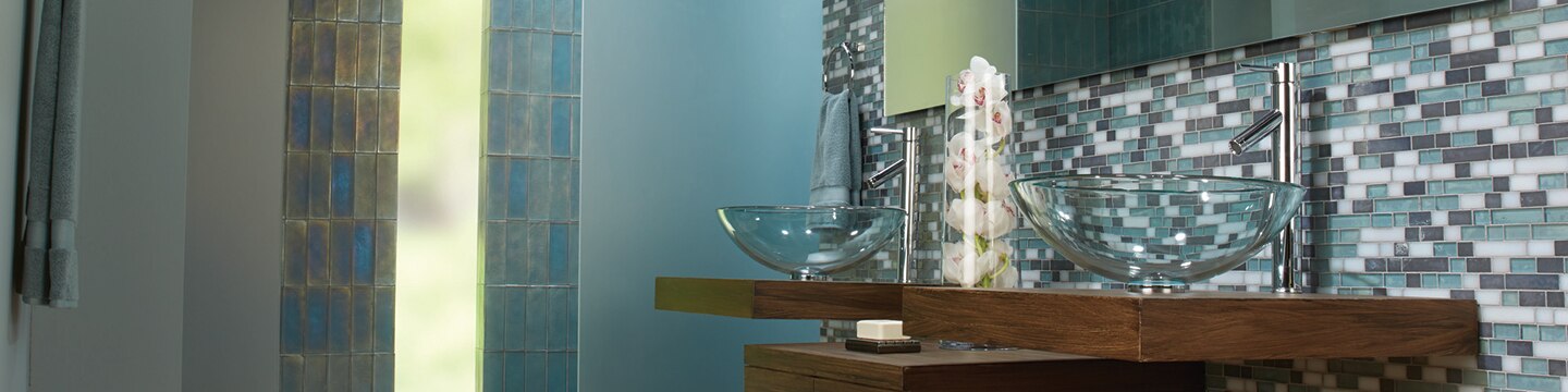 Spa-like bathroom with sea foam, white, and gray glass mosaic tile backsplash, butcherblock vanity with two top of counter glass sinks, open shower with blue glass tile wall and frosted glass door.