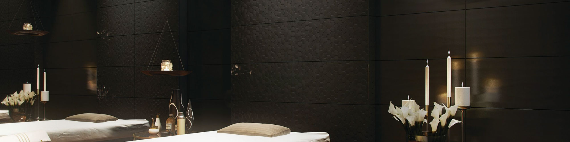 Spa massage room with black textured wall tile, massage table with blanket & pillow, floor tile that looks like wood planks, side tables with candles and flowers.