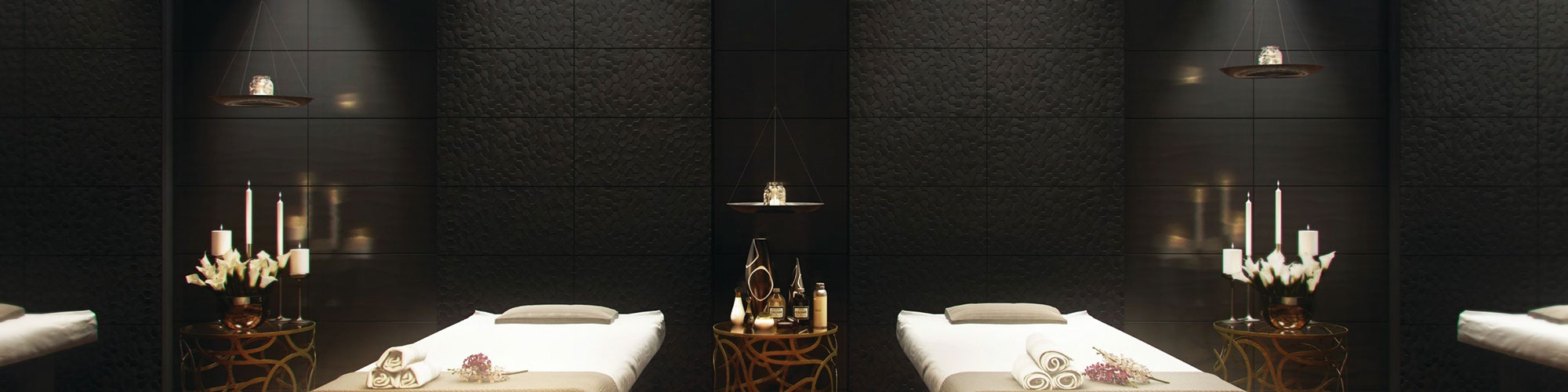 Spa massage room with black textured wall tile, massage tables with blankets & pillows, side tables with candles, flowers, and essential oils.