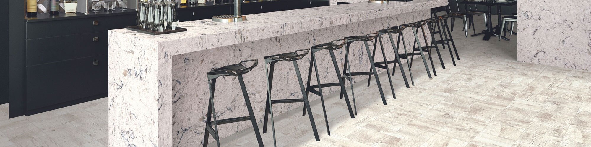 Hotel bar with floor tiling that looks like whitewashed wood, bar top of white quartz with black veining with a row of black bar stools.