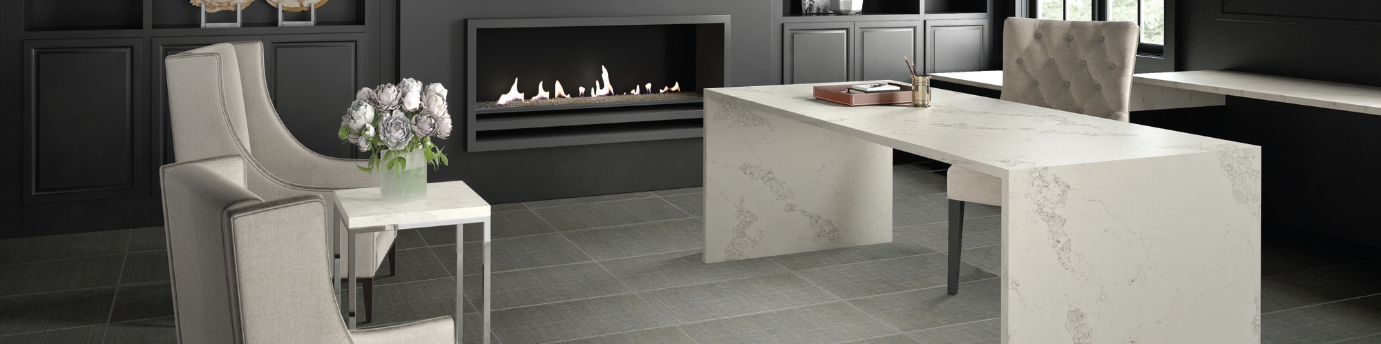 Home office with gray fabric look tile flooring, cream & gray quartz desk, two side chairs, and fireplace surrounded by dark gray built-in shelving.