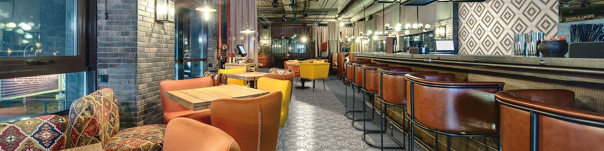 Restaurant with gray & beige encaustic floor tile, wood tables with bright mismatched chairs, encaustic wall tile, brick columns, and ceiling with exposed pipes & ductwork.
