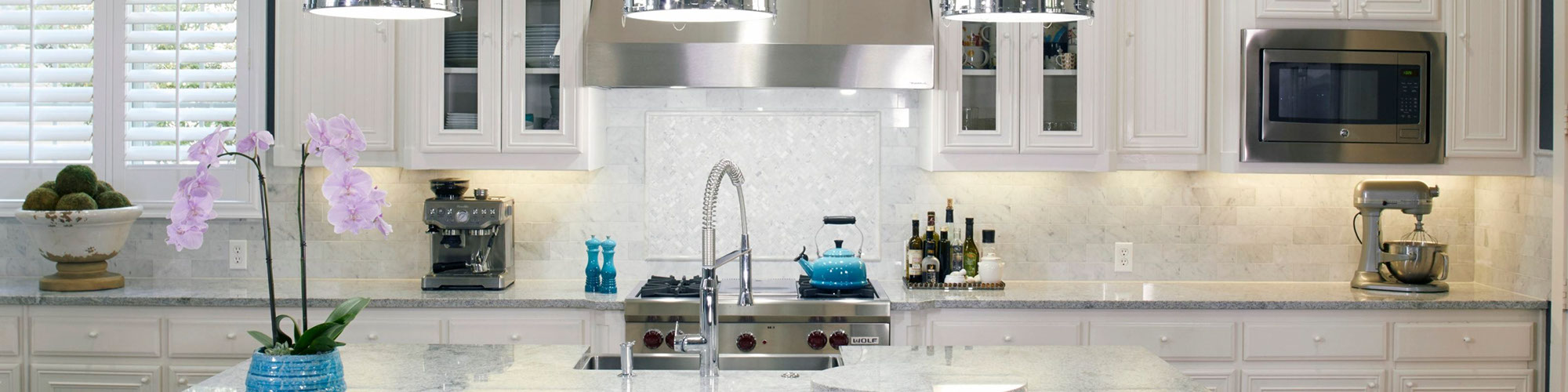 Kitchen with white granite countertop, island with sink, white marble backsplash with raised pencil trim outline and white cabinets.