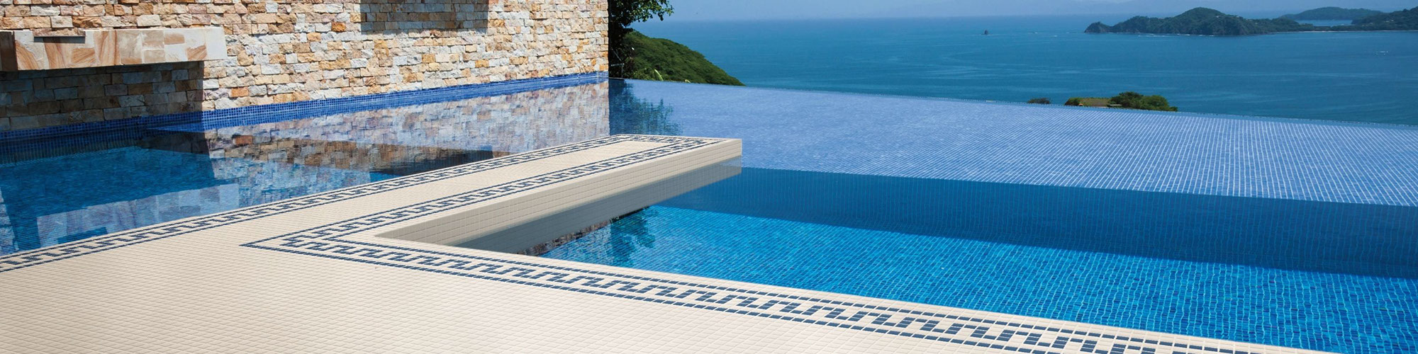 Infinity pool with white tile deck with blue accents and an ocean view with blue skies in the background.